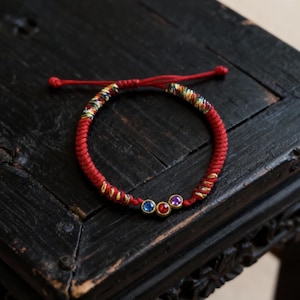 Könnte beinhalten: Rotes geflochtenes Armband mit drei kleinen, farbigen Edelsteinen. Das Armband hat ein gewebtes Design mit goldenen Akzenten und einem mehrfarbigen Abschnitt. Die Edelsteine sind blau, rot und lila.