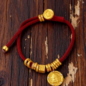 May include: A red cord bracelet with gold-colored accents. The bracelet features a series of gold rings, a square bead, and a circular pendant with a detailed design. The bracelet is adjustable and has a gold-colored clasp.