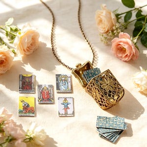 Könnte beinhalten: Ein goldfarbener Metall-Medaillon-Anhänger an einer Kette, gestaltet als aufklappbare Miniaturbox mit einem filigranen Blumenmuster. Im Inneren des geöffneten Medaillons befinden sich kleine rechteckige Karten mit blau gemusterten Rückseiten.