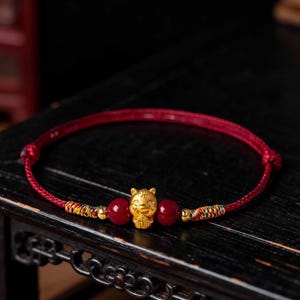 Könnte beinhalten: Rotes Armband mit einem goldfarbenen Katzenanhänger, zwei roten Perlen und goldenen Akzenten. Das Armband wird auf einer dunklen Holzoberfläche präsentiert, die die feinen Details hervorhebt.