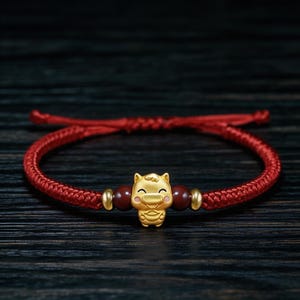 May include: Red braided bracelet with a gold-coloured pig charm. The bracelet has two dark red beads and gold-coloured accents. The bracelet is tied with a sliding knot. The bracelet's circumference is adjustable.