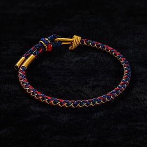 Puede incluir: Pulsera trenzada con hilos rojos, azules y verdes, acentuada con metal dorado. La pulsera presenta un cierre de barra en T y extremos de metal dorado. El diseño es intrincado y los colores son vibrantes sobre un fondo oscuro.
