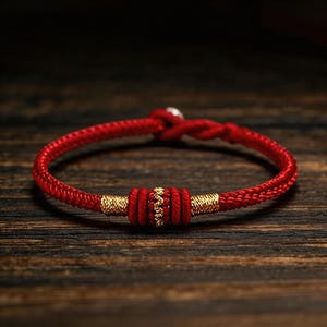 Op de afbeelding: Een rode gevlochten armband met goudkleurige accenten. De armband heeft een centraal decoratief element met goudkleurige kralen en twee goudkleurige banden aan weerszijden. Het wordt getoond op een donker houten oppervlak.