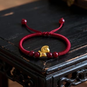 May include: Red braided bracelet with a gold horse charm and red beads. The bracelet is on a dark wooden surface. The horse charm is detailed with a small square design. The bracelet is tied with a red string.
