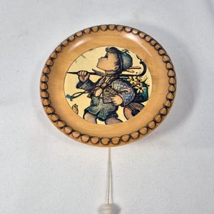 May include: A round, wooden decorative plate with a detailed border. The plate features a painted image of a young person carrying flowers and a musical instrument. A string with a small, white ball hangs from the bottom of the plate.