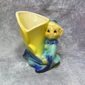 May include: A yellow and blue ceramic planter shaped like an elf holding a yellow flower. The elf has a blue and white hat and a blue and white outfit.