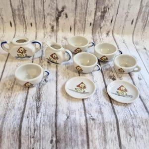 May include: A white ceramic tea set with a duck design. The set includes a teapot, creamer, cups, and saucers. The duck is yellow with a red hat. Blue accents are on the teapot and cups.