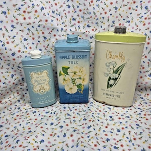 May include: Three vintage talc containers. One is light blue with the text "AVON Here's my Heart". Another is blue with "APPLE BLOSSOM TALC" and a floral design. The third is off-white with "Chambly Lily of the Valley Perfumed Talc".