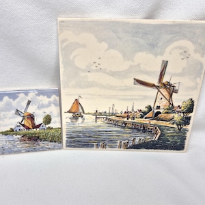 May include: Two ceramic tiles with Dutch windmill scenes. The larger tile depicts a windmill on a riverbank with a sailboat and town in the background. The smaller tile shows a similar windmill scene. Both tiles have a blue and white sky.