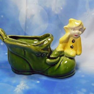 May include: A green ceramic boot planter with a yellow ceramic elf sitting on the side. The elf has a yellow hat and a yellow jacket with two black buttons.