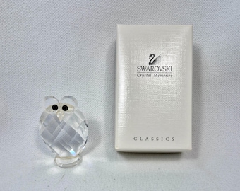 May include: A clear crystal owl figurine with black eyes and a small base. Beside the owl is a white box with the text "SWAROVSKI Crystal Memories CLASSICS". The owl is faceted, reflecting light.