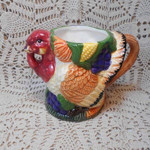 May include: A ceramic turkey-shaped mug with a detailed design. The mug features a red head, orange and brown feathers, and a green and yellow harvest-themed pattern around the rim. The handle is brown, and the interior is white.