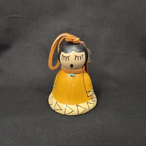 May include: A handcrafted Native American-style bell figurine. The bell is painted with a yellow dress, black hair, and a turquoise bead necklace. The base features a geometric design, and a leather loop is attached for hanging.