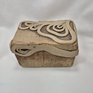 May include: A small, rectangular ceramic box with a textured, beige surface. The lid and sides feature raised, swirling, cream-colored designs. The box has a rounded edge and a lid that fits snugly.