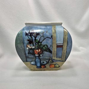 May include: A flat, rounded ceramic vase featuring a painted still life scene of a blue vase with flowers, a plate, and fruit on a table. The background depicts a window with blue and beige tones. The rim of the vase is finished with a gold trim.