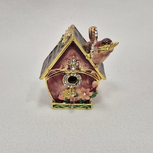 May include: A decorative birdhouse-shaped trinket box. The box is burgundy with gold trim and a small bird perched on the roof. The birdhouse is adorned with rhinestones and floral accents. The bird is brown with a yellow beak.