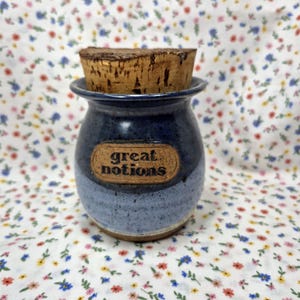 May include: A small, blue ceramic jar with a cork stopper. The jar has a label that reads "great notions" in a brown, oval-shaped label. The jar is a mix of dark and light blue colors.