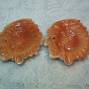 May include: Two ceramic ashtrays shaped like Native American chiefs. The ashtrays are a glossy, light brown color with detailed facial features and headdresses. The ashtrays are placed side by side on a white lace surface.