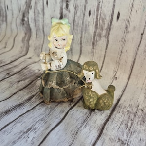 May include: A vintage porcelain figurine of a girl with blonde hair and a green bow, seated and holding a leash attached to a poodle. The poodle is green and white. The girl is wearing a green dress with lace trim.
