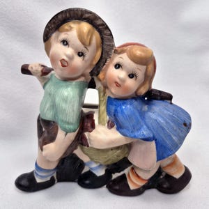 May include: A vintage ceramic figurine depicting a boy and girl. The boy wears a green shirt, brown shorts, and a hat. The girl is in a blue dress and a red hair ribbon. They appear to be carrying a basket. The figurine is painted in soft pastel colors.