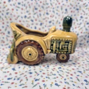 May include: A ceramic tractor planter in shades of tan, brown, and green. The planter has a detailed design, including wheels, a seat, and a smokestack. The tractor is a decorative item for a home or garden.