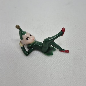 May include: A small figurine of an elf lying on its side, wearing a dark green outfit with a matching pointed hat topped with a gold-colored tip. The elf has a pale face, dark eyelashes, and red shoes, posed with one leg bent upward.