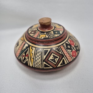 May include: A small, round, ceramic jar with a lid. The jar is painted with geometric patterns in black, red, yellow, and brown. The lid has a small, cork knob. The jar is likely used for storing small items.