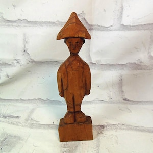 May include: A hand-carved wooden figurine of a person wearing a hat and overalls. The figure is standing on a square base. The wood has a warm, brown tone, and the carving details are visible.