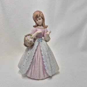 May include: A porcelain figurine of a person with light brown hair wearing a pink dress with a white, textured, open-front overskirt. The figure holds a small brown basket filled with light-colored flowers and features a small blue bow at the waist.