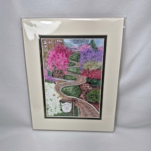 May include: A framed artwork depicting a winding stone path through a colorful garden. The path is bordered by vibrant pink, purple, and red flowering trees and bushes. A sign with text is visible near the path. The artwork is matted and framed.
