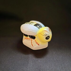 May include: A ceramic bee figurine with a yellow head and body, white wings, and black and yellow stripes. The bee has a smiling face with a black eye and red legs. The figurine is on a dark background.
