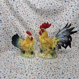 May include: Two ceramic rooster figurines. The roosters have yellow bodies, red combs, and black and blue tail feathers. They are standing on a base of green grass. The background is a white floral pattern.