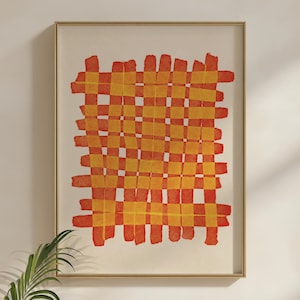 May include: Abstract art print featuring a woven pattern in shades of orange and yellow. The artwork is framed in a gold-colored frame and hangs on a white wall. The design has a grid-like structure with a textured, watercolor-like appearance.