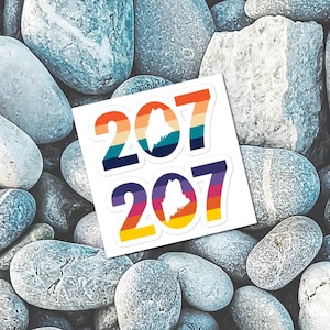 Op de afbeelding: Twee stickers met het nummer "207" in een retro lettertype, met een kleurrijk gestreept ontwerp. De staat Maine staat in het midden van de nul. De stickers liggen op een achtergrond van grijze en witte stenen.