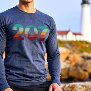 Puede incluir: Una camiseta azul marino de manga larga con el número "207" en un esquema de colores retro. El diseño incluye una silueta del estado de Maine. Un faro y un paisaje costero están en el fondo.