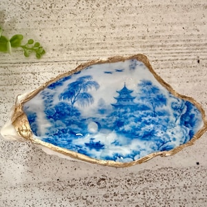 May include: A decorative oyster shell dish with a blue and white Asian-inspired landscape design. The shell's edge is trimmed with gold paint, creating a unique and elegant accent. The dish is ideal for holding small items.