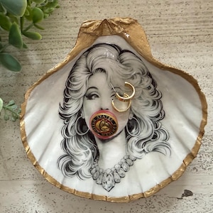 May include: A decorative seashell trinket dish with a gold-painted rim. The dish features a black and white illustration of a woman with gold hoop earrings and a pink object in her mouth. A necklace is also visible.