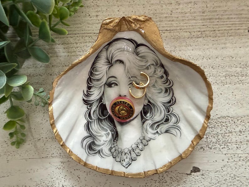 May include: A decorative seashell trinket dish with a gold-painted rim. The dish features a black and white illustration of a woman with gold hoop earrings and a pink object in her mouth. A necklace is also visible.