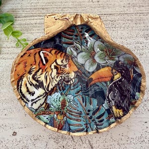 May include: A decorative seashell with a gold-painted rim. The interior features a vibrant illustration of a tiger and a toucan amidst tropical foliage. The artwork is set against a teal background with white flowers.
