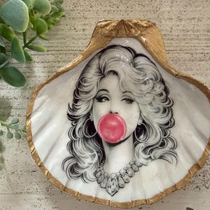 May include: A decorative seashell with a gold-painted edge. The shell features a black and white illustration of a woman blowing a bubblegum bubble. The woman is wearing large hoop earrings and a jeweled necklace.