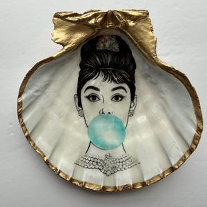 May include: A decorative seashell with a gold-painted edge and a black and white illustration of a woman blowing a bubble. The woman has dark hair and is wearing earrings and a necklace. The bubble is a light blue color.