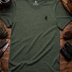 May include: A heathered olive green short-sleeve t-shirt with a small black embroidered silhouette of a creature on the left chest. The inner neck label features the text Squatch Outfitters with a small graphic of the same creature.