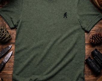 May include: A heathered olive green short-sleeve t-shirt featuring a small black embroidered silhouette of a Bigfoot on the left chest. The inner neck label displays the text Squatch Outfitters with a small Bigfoot icon above the brand name.