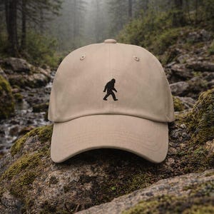 May include: A tan baseball cap with a black silhouette of Bigfoot embroidered on the front. The cap is set against a backdrop of a forest and moss-covered rocks.
