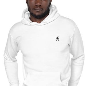 May include: A white long-sleeve hooded sweatshirt featuring a small black embroidered silhouette of a Bigfoot figure on the left chest. The garment includes a drawstring hood with silver-toned metal eyelets and a large front kangaroo pocket.