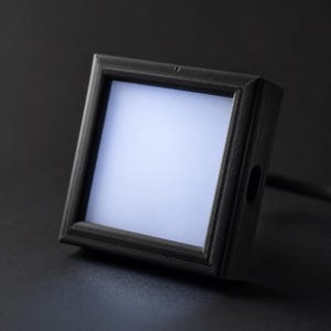 Puede incluir: Un marco cuadrado negro con una superficie texturizada y un centro blanco brillante iluminado. El marco tiene un pequeño agujero en el lateral y está sobre un fondo oscuro. El objeto parece ser una caja de luz o una pantalla.