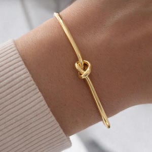 May include: A gold-colored bangle bracelet featuring a knot design at the center. The bracelet is a simple, elegant piece of jewelry. The smooth, polished surface reflects light, highlighting the warm tone of the metal. The bracelet is worn on a wrist.