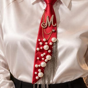 May include: A red satin necktie featuring a large rhinestone-encrusted letter M, gold-tone chains, scattered white faux pearls of varying sizes, and a cascading fringe of silver-tone metal chains attached to the side of the fabric.
