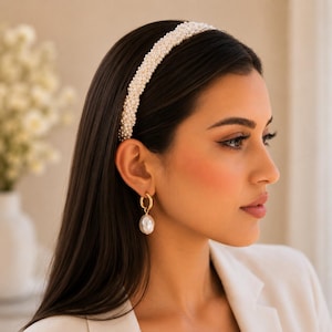May include: A wide headband densely covered in small, round, white faux pearls. The headband is worn with a gold hoop earring featuring a single, large, teardrop-shaped white pearl pendant that hangs from the bottom of the circular gold metal hoop.