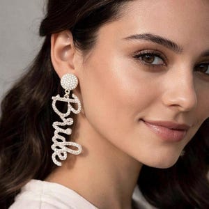 May include: A sparkling, dangling earring with the word "Bride" spelled out in cursive script. The earring is made of small, clear crystals and has a round, crystal-covered stud at the top. The jewelry is a bridal accessory.